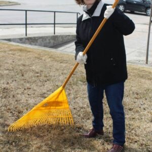 woman holding. a yellow rake on grass with a parking lot and cars in the backgroud