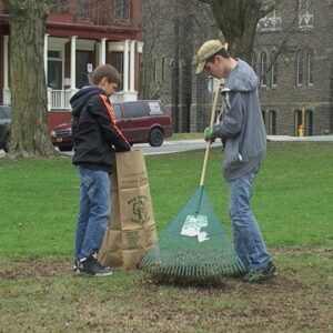 two boys working together with a rake and bag cleaning up grassy area with buildings in the background